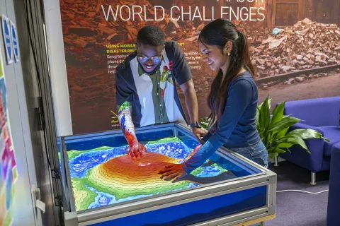 Two students exploring an interactive topographic sandbox display with projected contour and elevation colours.