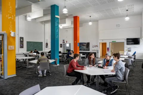A study and social space on a university campus. In the foreground, there is a group of four students sat around a table. There are more tables with students in the background, and the pillars around the room have mathematical equations written on them.