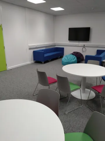 An empty room with sofas, beanbags and round tables with chairs surrounding them. There is also a TV mounted on the wall.