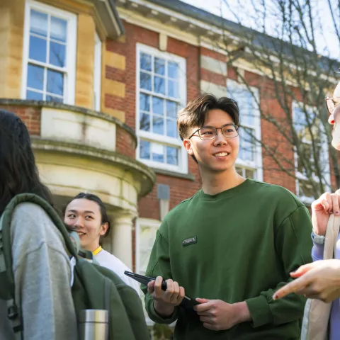 Two postgraduate students smiling at each other while walking outside the Parkes Building, also known as Building 65, on the University of Southampton's Avenue Campus. They are part of a larger group of students.