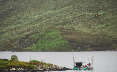 A drone hovers over a lake near a small floating platform with equipment, surrounded by lush green hills. The scene suggests a scientific or environmental study in a remote natural location.