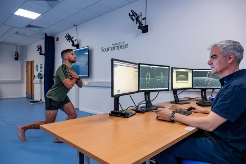 A man performs a lunging squat with motion capture markers, while another man monitors data on screens in a lab.