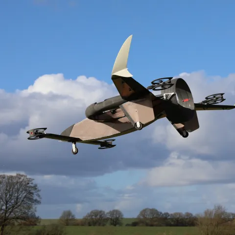 A drone with vertical stabilisers and multiple rotors flying through a blue sky with scattered clouds, above a green field dotted with trees.
