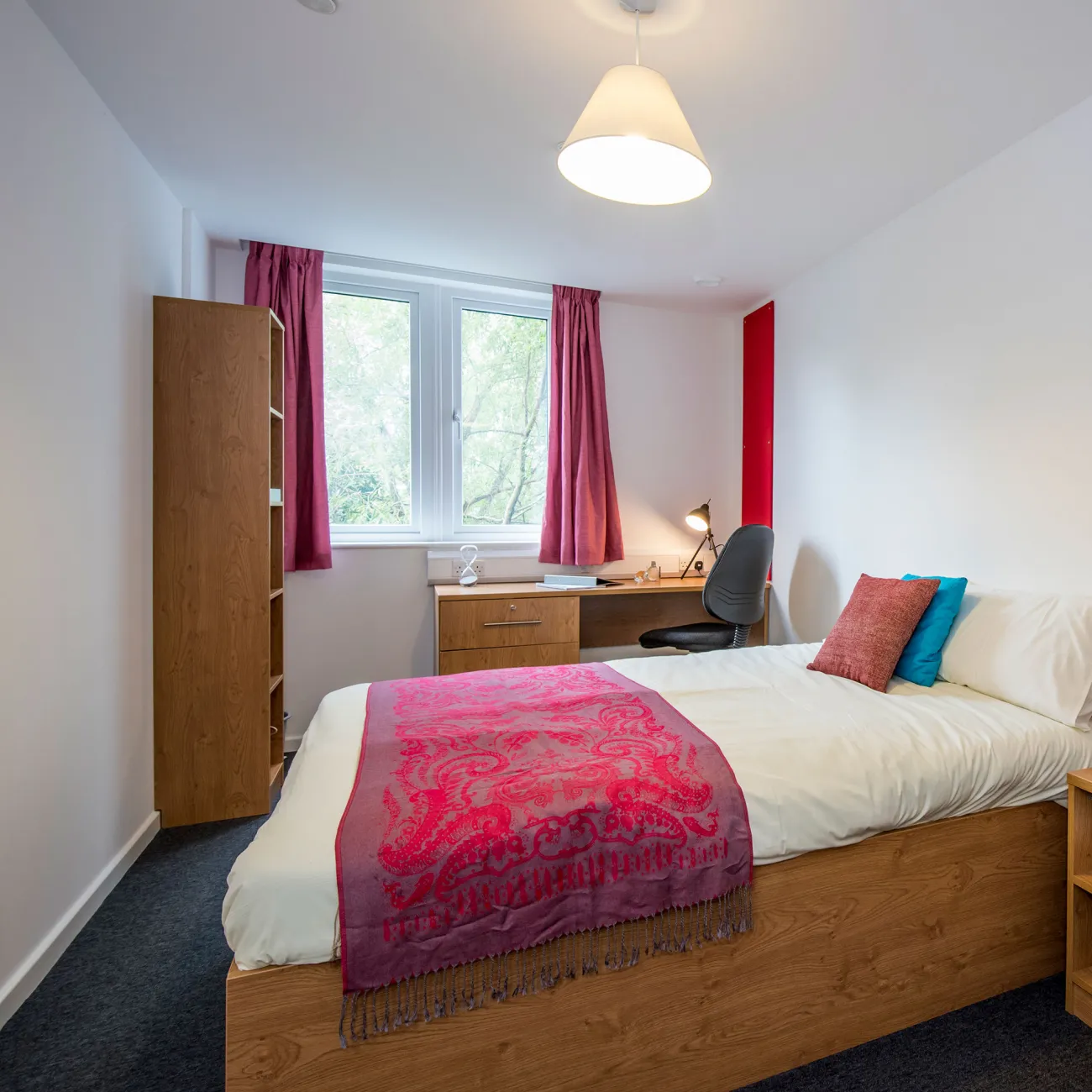 Bedroom with a neatly made double bed and nightstand. A wooden desk and open window are visible in the background.