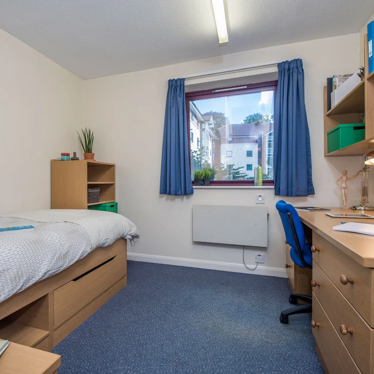 A window lets light into a bedroom where a neatly made bed and desk can be seen.