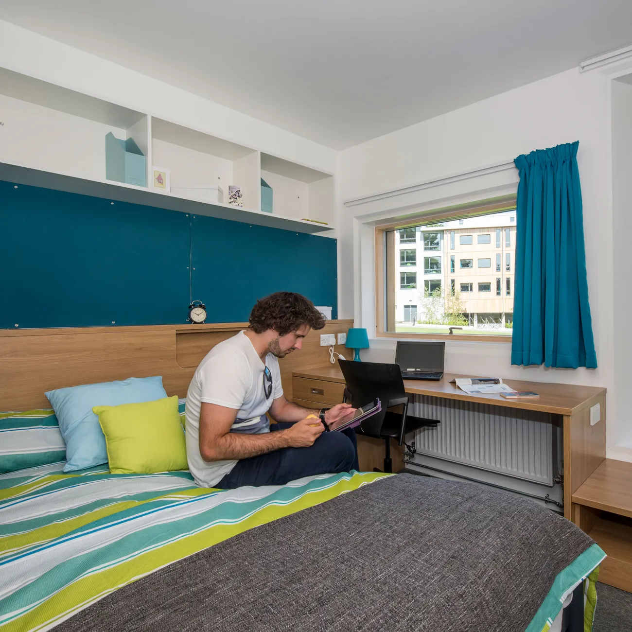 A student sits using a tablet on a large, brightly coloured bed in a modern bedroom. Modern buildings and a grassy courtyard can be seen through the windows.