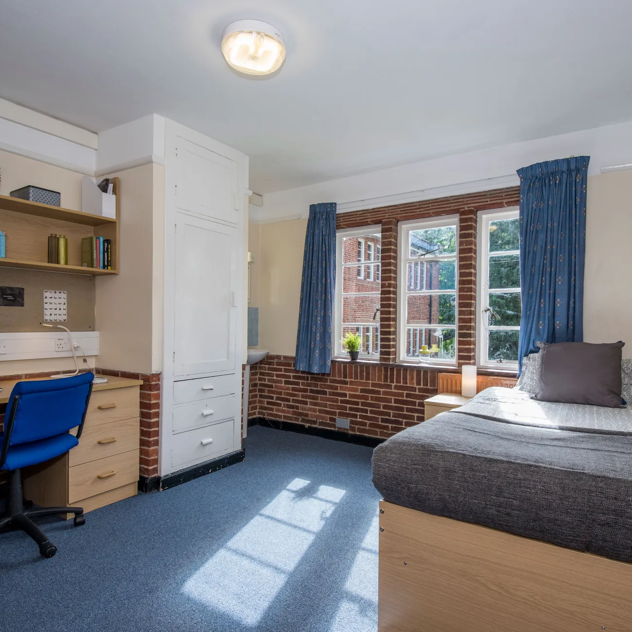 Light from a large window fills a spacious bedroom with desk, chair and neatly made bed.