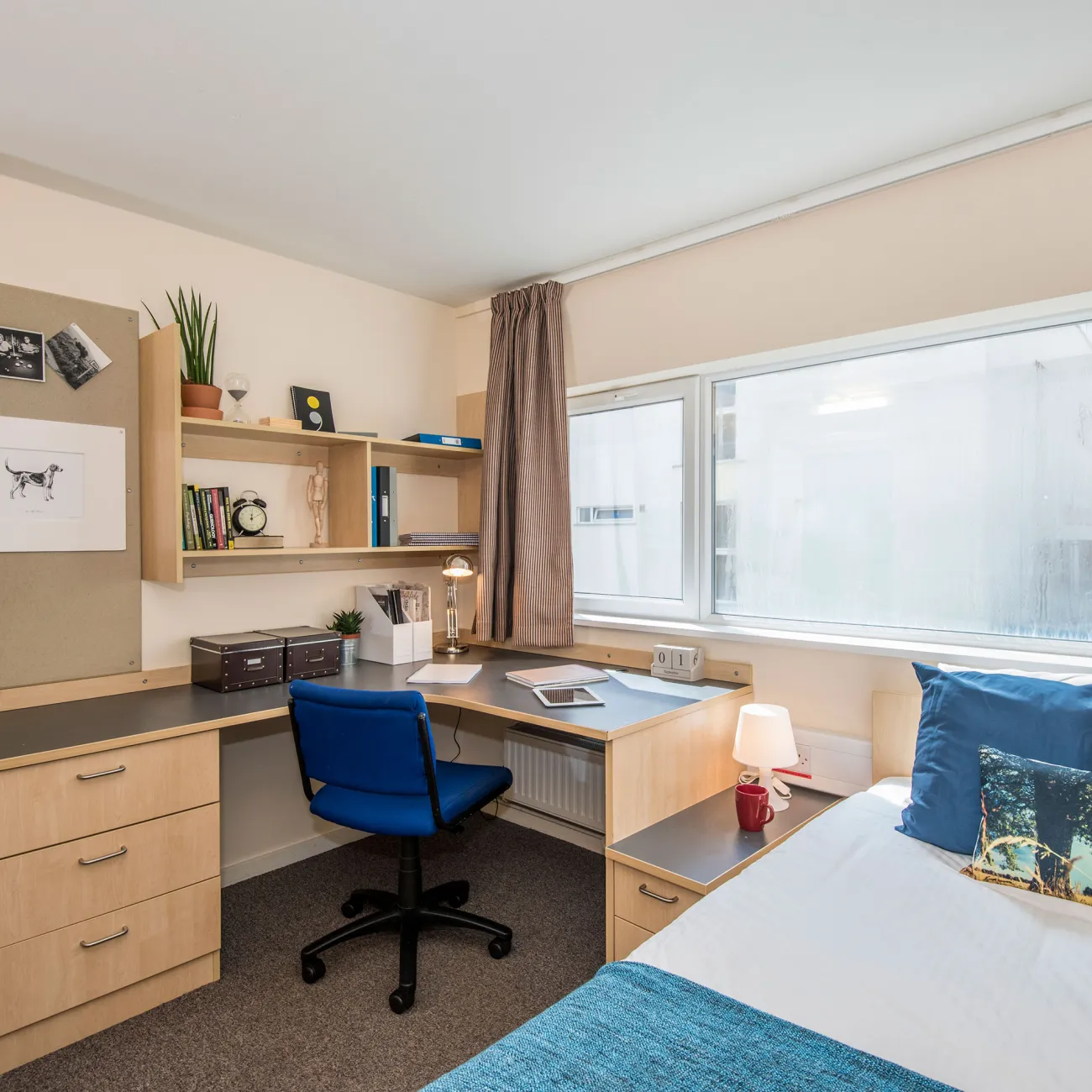 A large window lets light into a bedroom where a neatly made double bed, wash basin and desk can be seen.