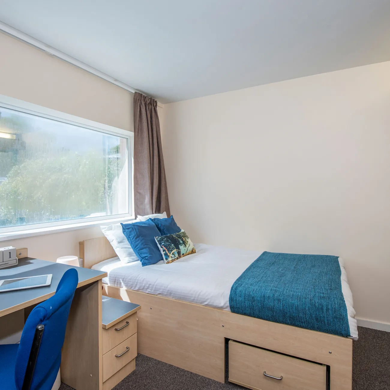 A large window lets light into a bedroom where a neatly made double bed, wash basin and desk can be seen.