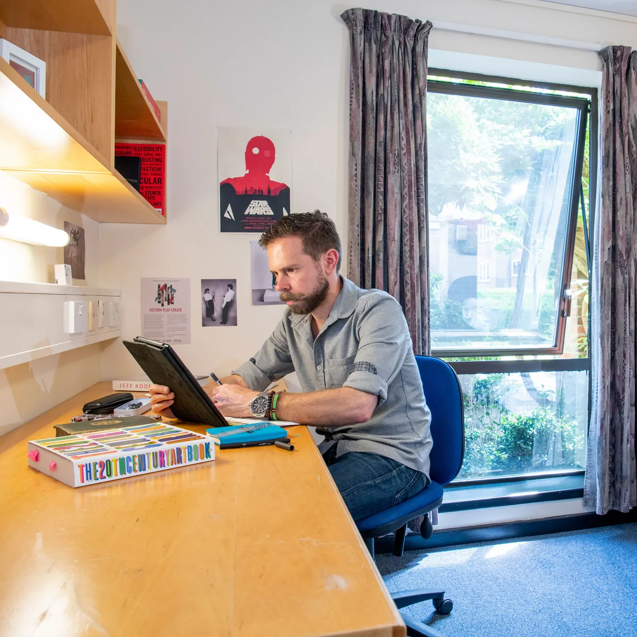 A student sits looking at a tablet computer while working at a desk. A bed and wardrobe can be seen behind him, an open window lets light into the room.