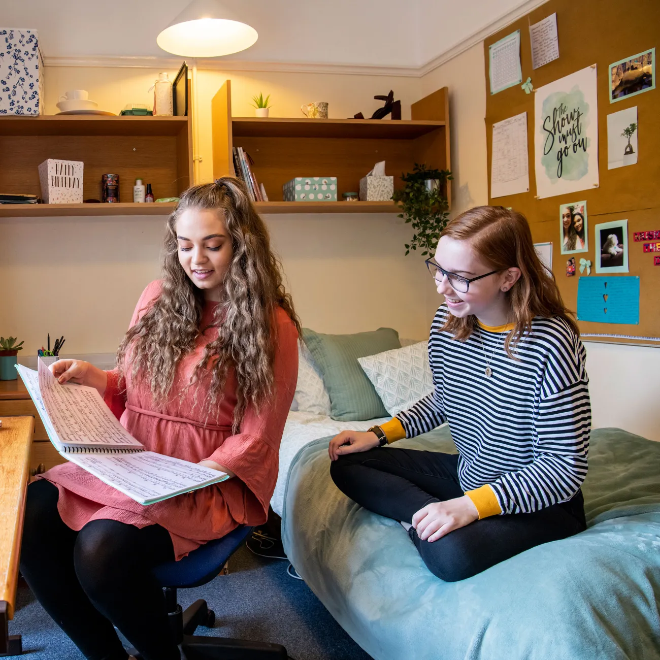 Two students sat in a bedroom talking and looking at books and notes. Light streams in through a large window.