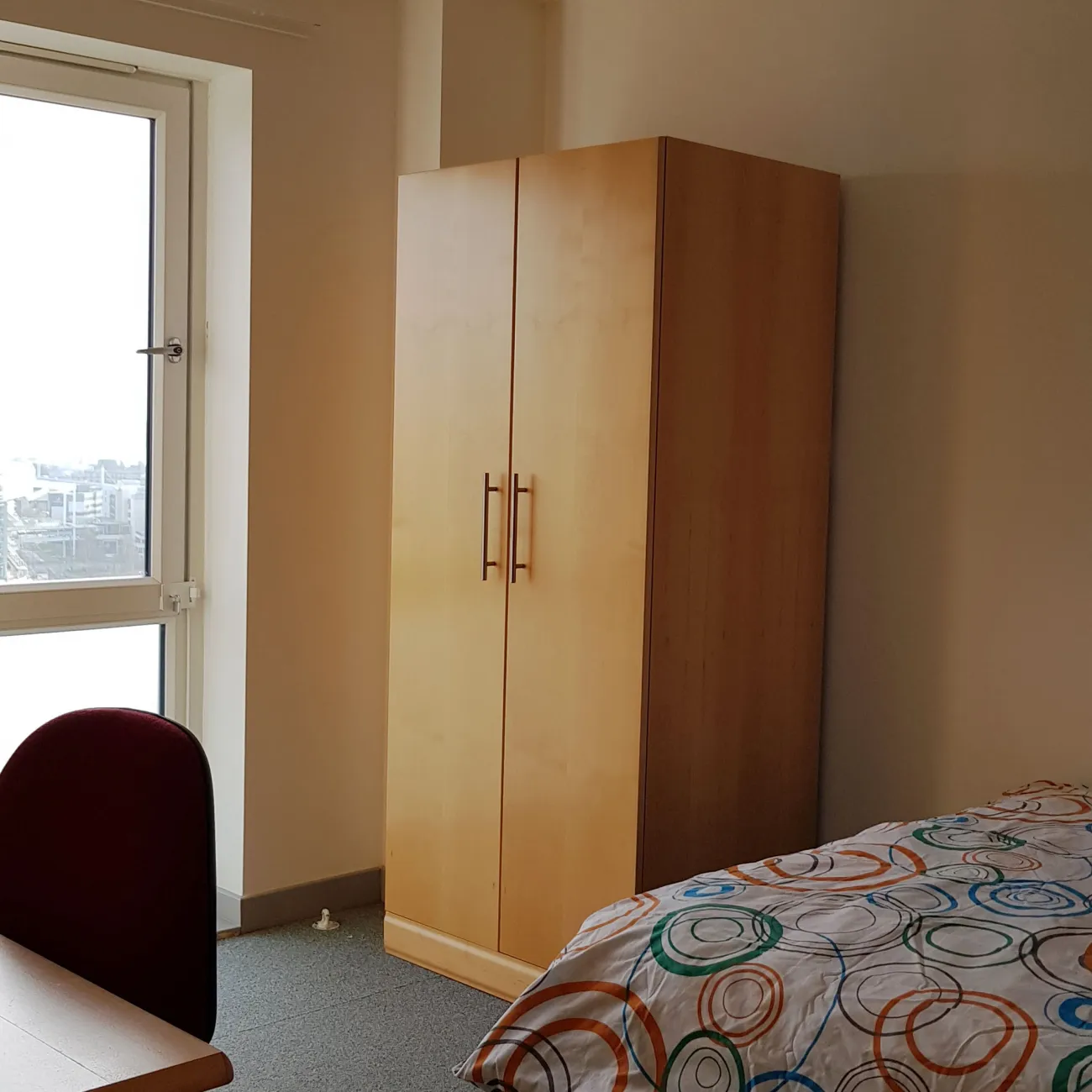 View of a bedroom showing a double bed with floral sheets, a large wooden wardrobe and a desk. A large window lets light into the room.