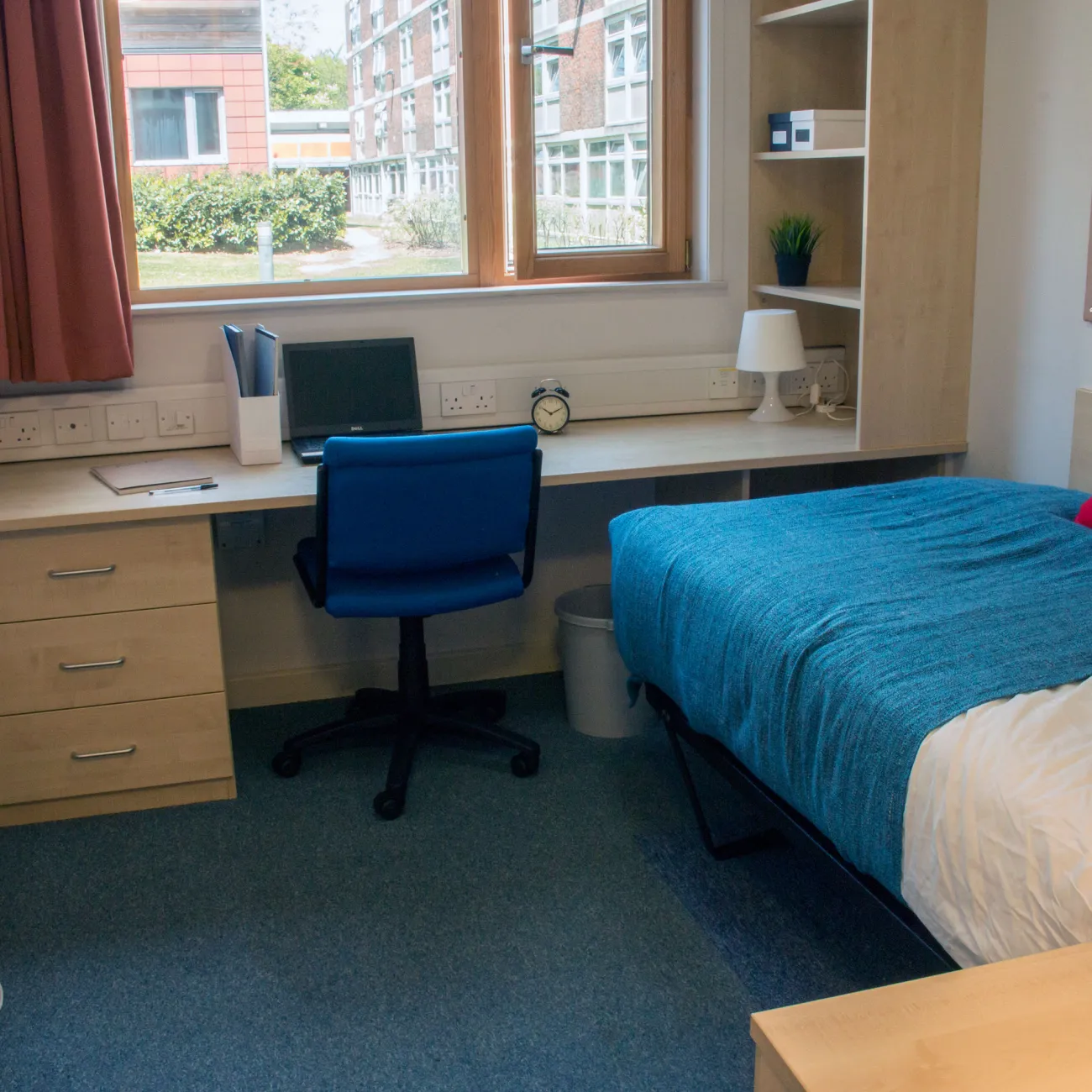 An empty student bedroom containing a neatly made single bed and a large desk with chair, beneath a window.