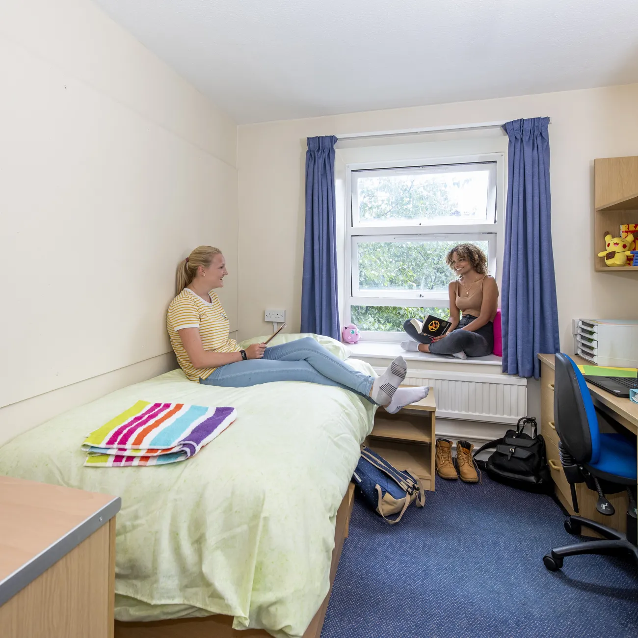 Two students chatting in a bedroom. One sits browsing with her tablet on a single bed while the other looks up from their book, sat on the windowsill.