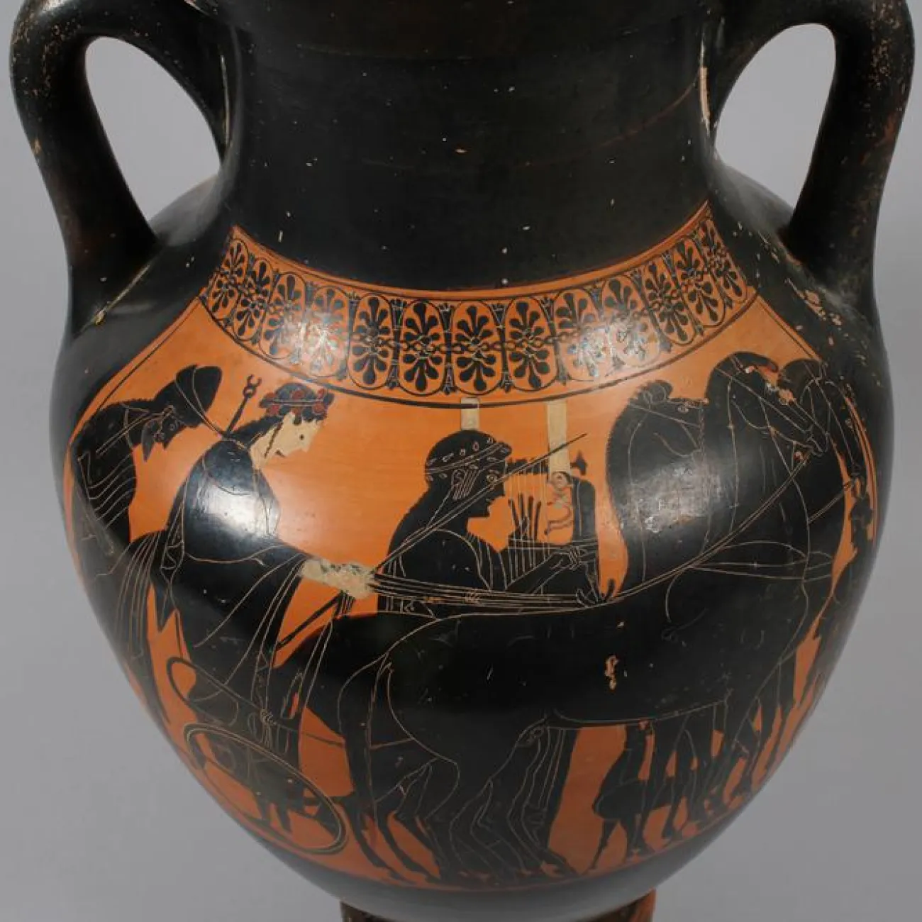 A black and orange amphora pot. The artwork on the side shows three people, one of them are holding the reins of three horses.
