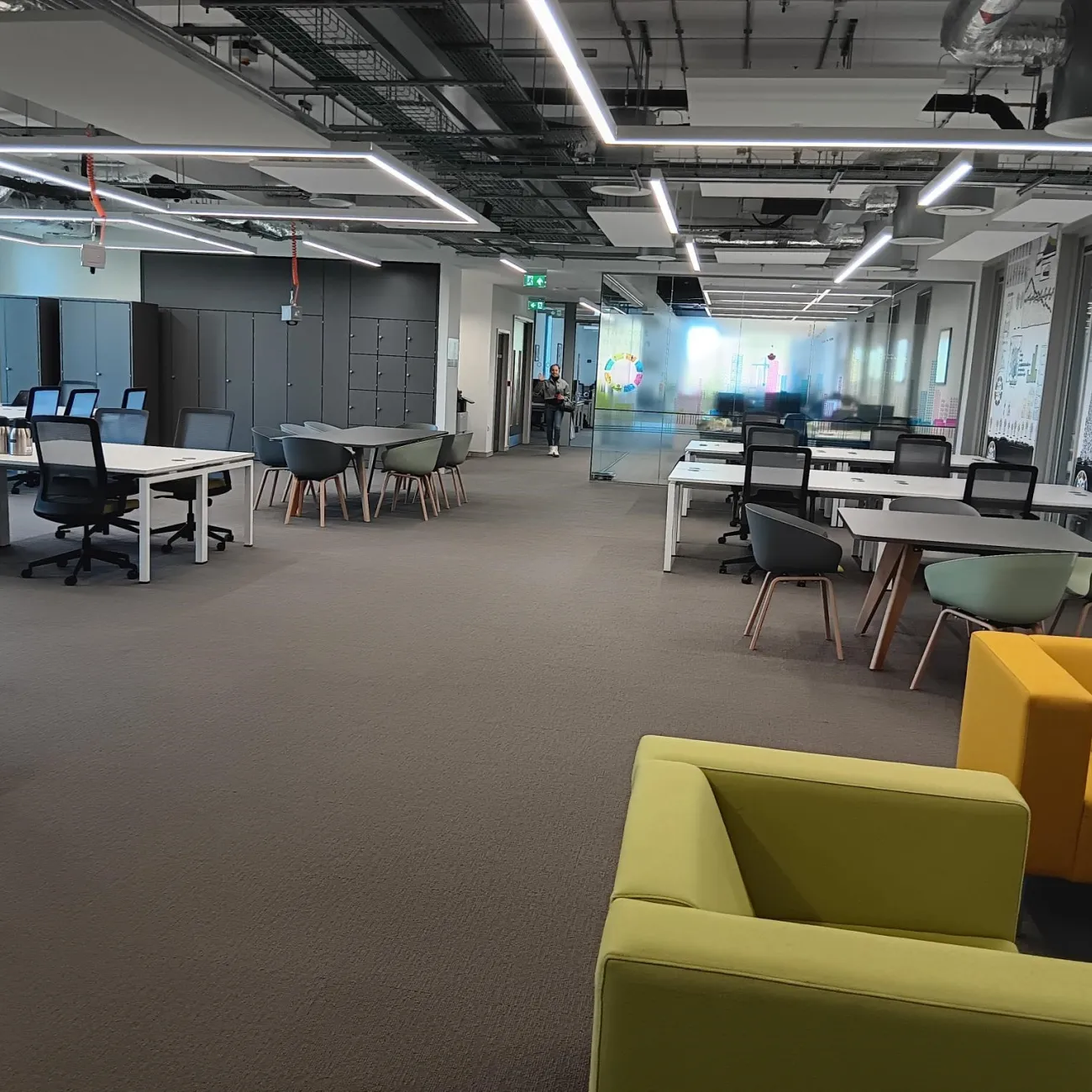 A modern open-plan office with white desks, black chairs, and grey lockers. Bright yellow and green lounge chairs are in the foreground.