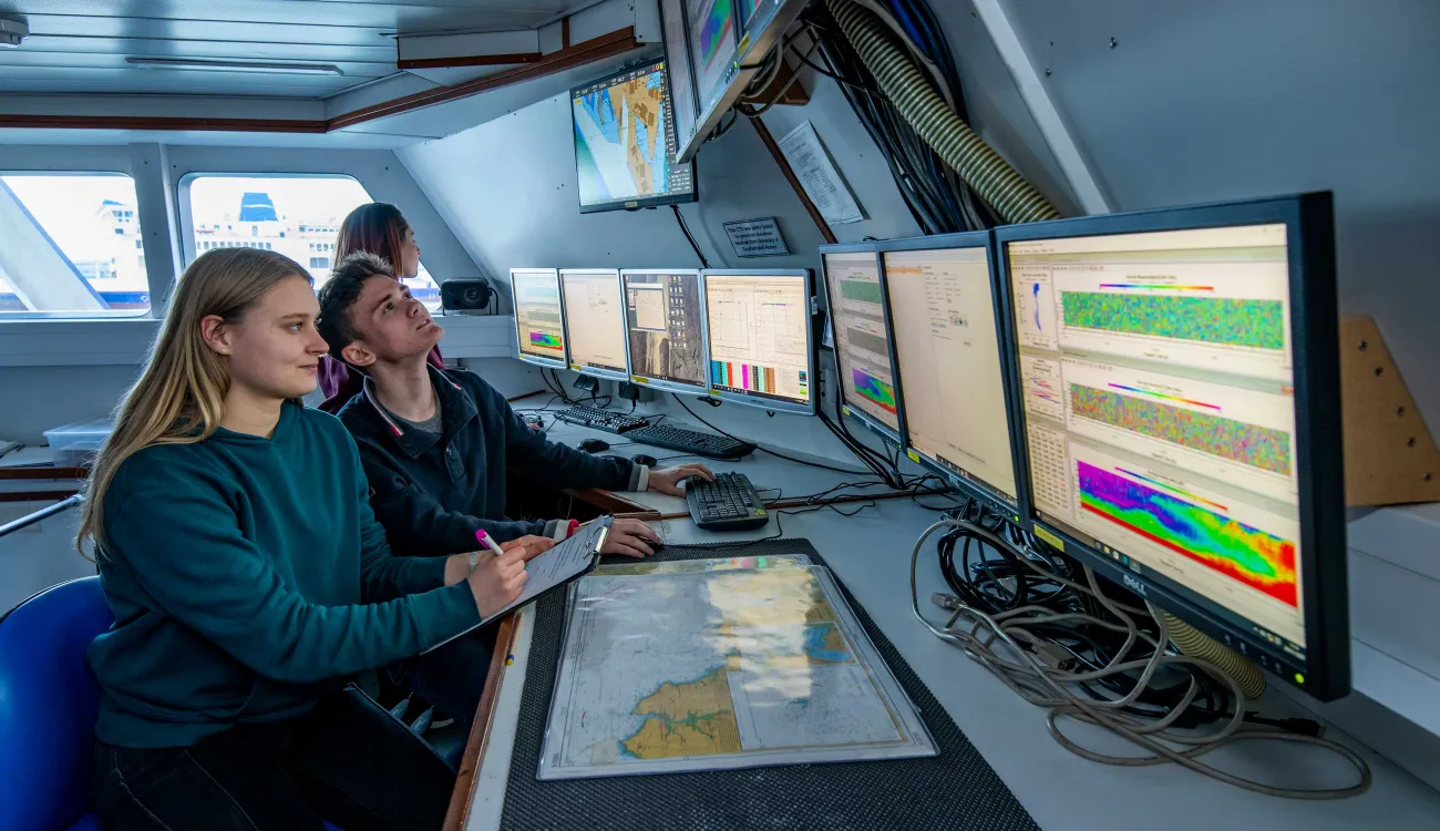 Two students sit at a desk inside a room on a boat. They observe a bank of computer screens displaying various data and information.