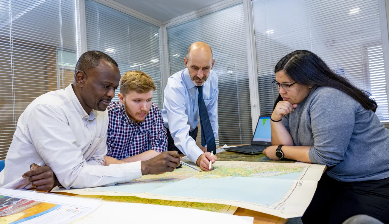A Southampton Geospatial researcher talks with team members, standing over a map, with a laptop with a data visualisation to the side