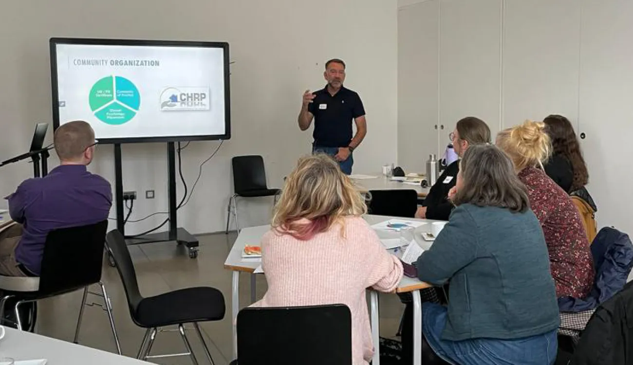 Professor Nick Maguire speaks at a community research impact event. A group of 6 adults sit around desks, listening, as Nick talks animatedly