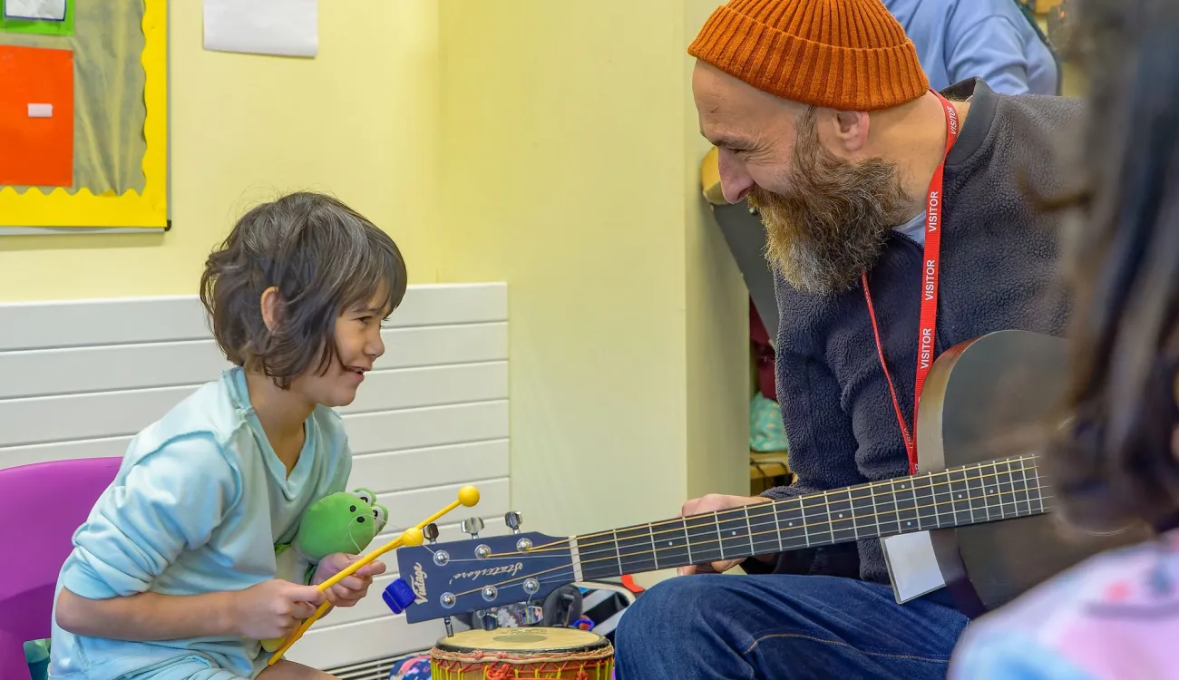 A bearded man wearing an orange knit hat and visitor lanyard plays guitar while interacting with a young child in a light blue shirt who is holding drumsticks. They appear to be in a colorful classroom or community center setting, engaged in a musical activity together.