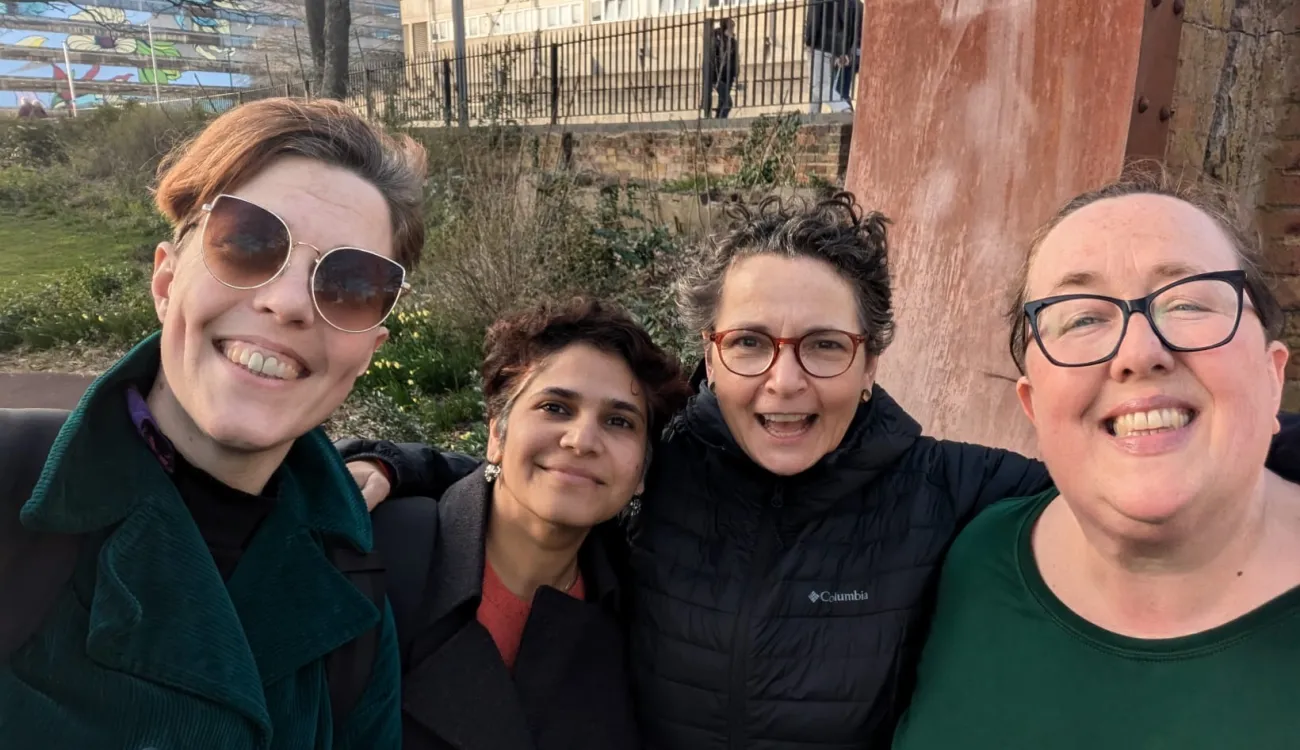 Four women pose together for a cheerful group selfie outdoors, all smiling warmly at the camera. They are dressed in winter clothing including coats and jackets, with urban buildings and greenery visible in the background.