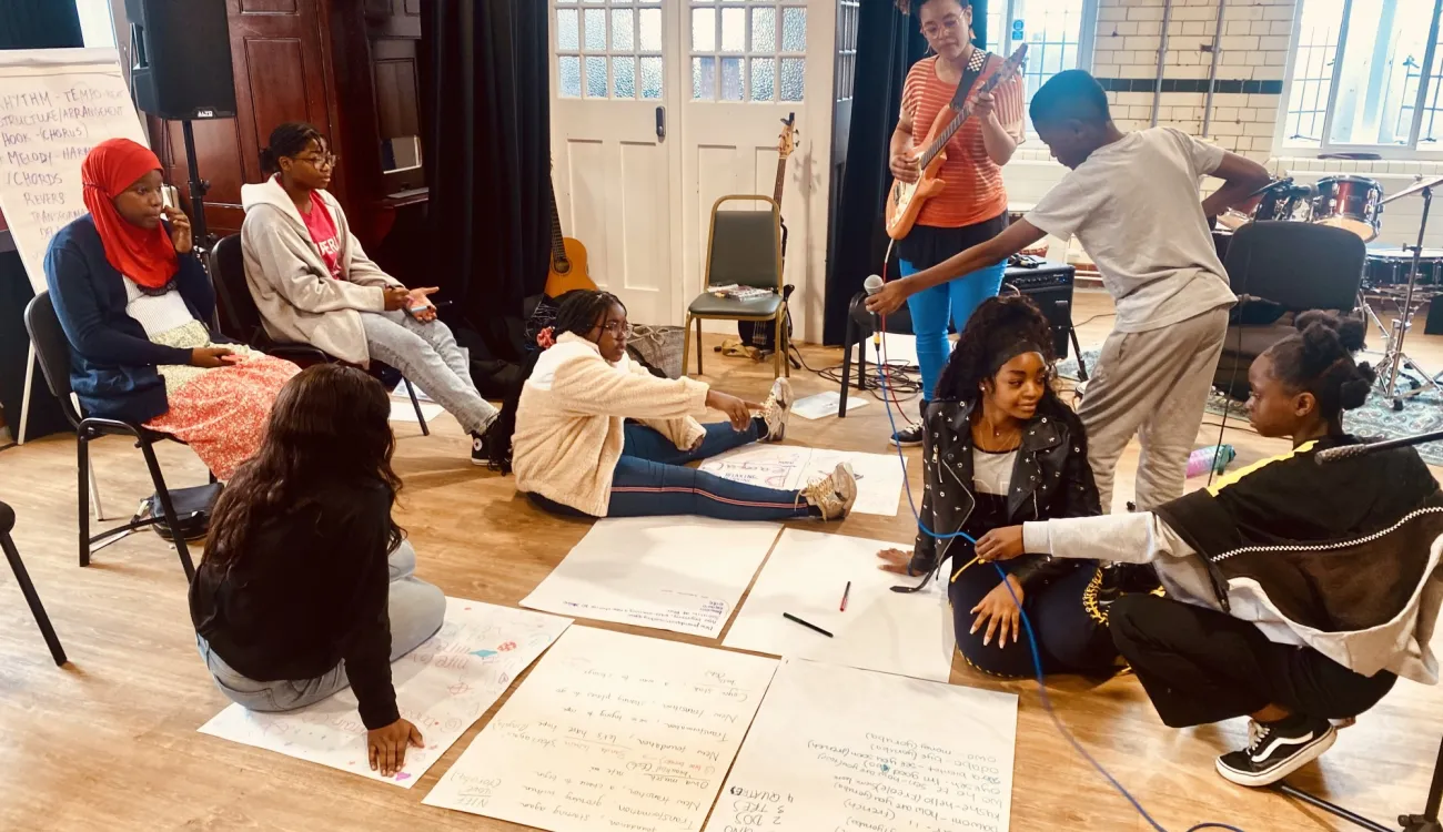 A group of young people participate in a creative workshop or music session in a large room with wooden floors, surrounded by large sheets of paper with writing and musical instruments including guitars and drums. Some participants are seated on chairs while others sit on the floor, with one person playing guitar and recording equipment visible in the scene.Retry