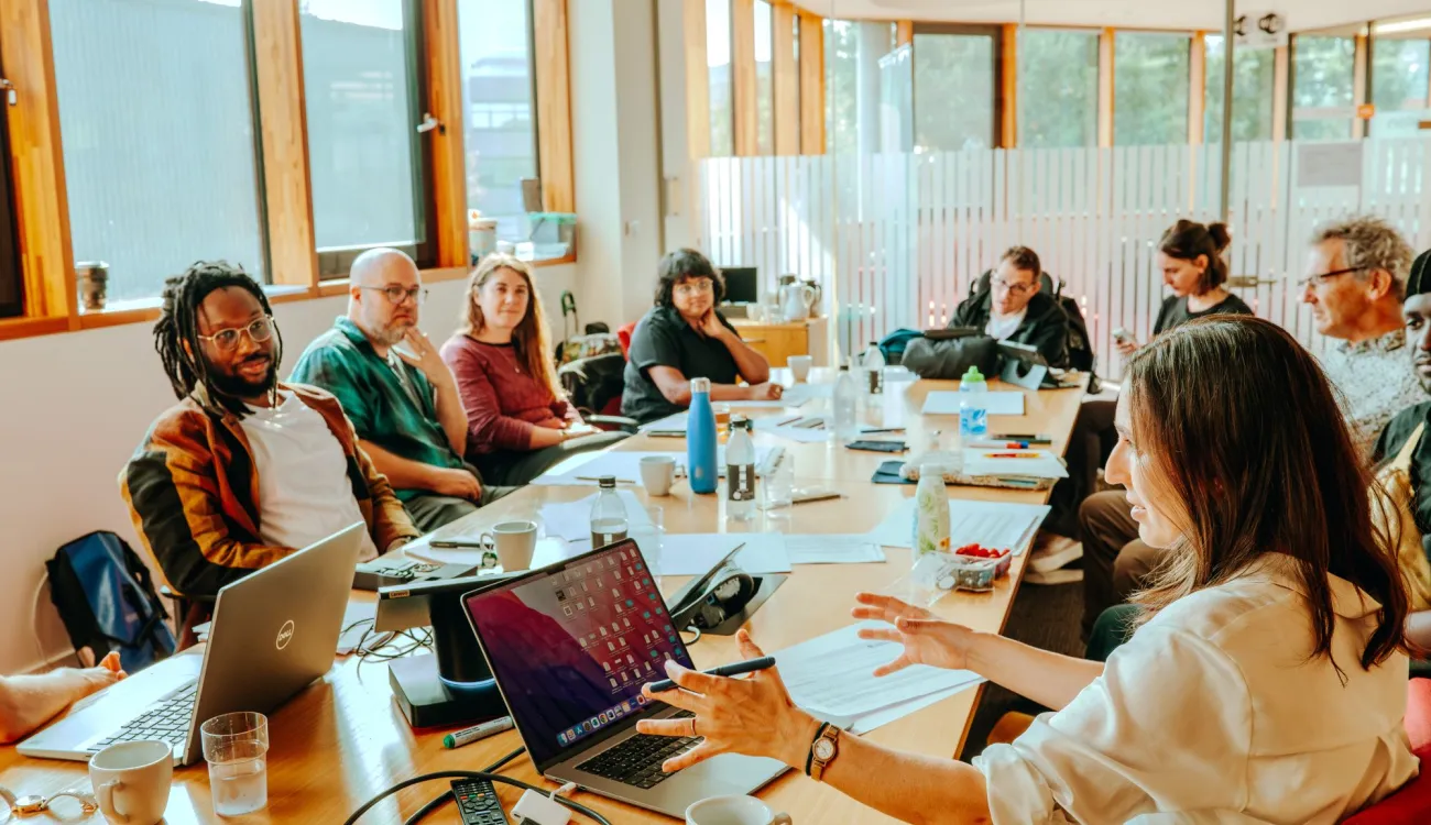 A diverse group of people sit around a large wooden conference table in a bright, modern meeting room with floor-to-ceiling windows, engaged in discussion with laptops, notebooks, and coffee cups spread across the table. The natural lighting and contemporary architecture create a collaborative workspace atmosphere for what appears to be a business meeting or workshop.