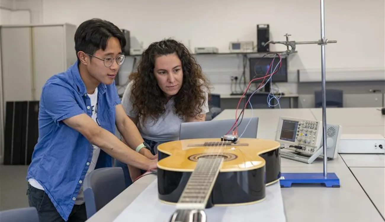 Two researchers, a man and a woman, prepare a vibration measurement setup on a classical guitar placed on a laboratory table.