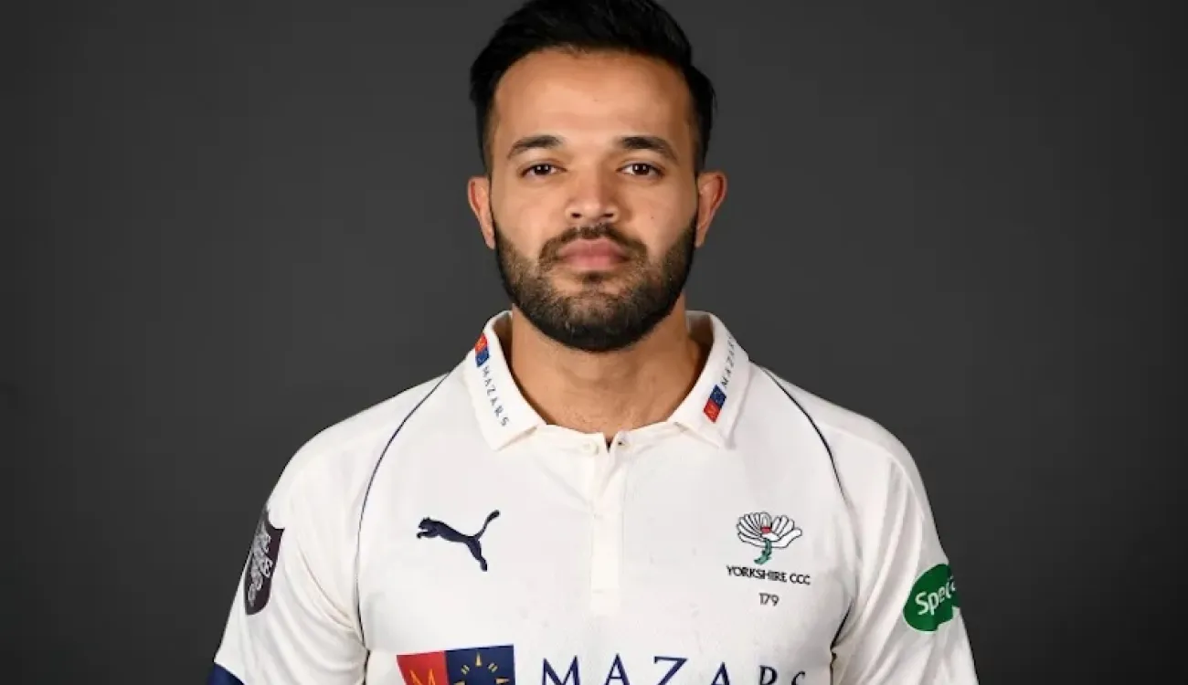 Azeem Rafiq in Yorkshire cricket shirt, facing camera against a dark background.
