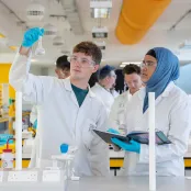 Two students wearing lab coats and protective glasses conduct an experiment. One holds up a glass beaker, while the other takes notes. They are in a laboratory, surrounded by a variety of chemistry equipment.