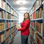 A student smiling over her shoulder as she carries a pile of books through the stacks in Hartley library.
