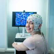 A student sits in front of a computer and turns to smile towards the camera. She is wearing a head cap that is connected to electrodes to monitor brain activity.