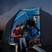 A small, domed rooftop observatory opens onto a starry night sky. Inside the observatory, two students use a telescope and a laptop to collect data.