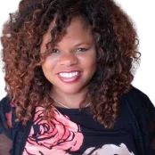 A photo of public contributor, Traci Carroll. She is wearing a black top with oversized pink and white roses. She has beautiful curly natural hair.