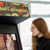 Laura Alford, BA Games Design and Art student looking at an arcade game console named Morbid Curiosities. There is a child like doll on the screen and 2 candle sticks on the game console. 