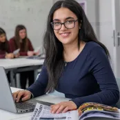 Head and shoulders portrait image of Seerat Chatha, BA Film, TV and Digital Media student smiling at the camera. She has her hands on a laptop and a magazine on the desk in front of her.