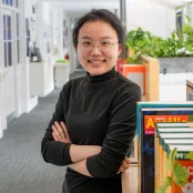 Head and shoulders portrait image of Chang Gao, MA Translation and Professional Communication Skills student smiling and looking at the camera. She is standing with her arms crossed next to a cupboard in a corridor.