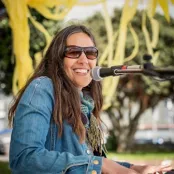 Emily Macklow is sitting playing a piano and has a microphone in front of her face.  She is smiling and looking at the camera.