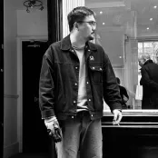 A black and white image of Linhao Li standing outside the glass front of a shop. There are people browsing inside the shop.