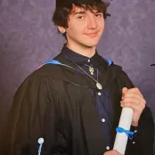 Samual Marsh dressed in his graduation robes and mortar board, holding a paper degree rolled up and tied with a blue ribbon.
