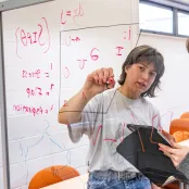 Jodie Adams is writing an equation on a vertical clear board with a red pen.
