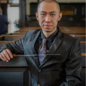 Xinda Zhu is sitting in an auditorium, looking at the camera and holding a baton.