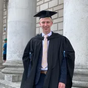 Health Psychology masters student, Nathan is dressed in his graduation robes standing outside a white stonebuilding with columns.