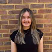 Portrait image of Law student Maddy Nicholl smiling at the camera. There is a red brick wall behind her. 