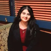 MSc Data and Decision Analytics student, Marwa Ashfaq is sitting down smiling at the camera. There is a closed window blind behind her.