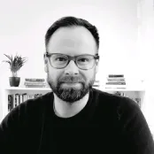 Black and white portrait headshot of a man. He wears a black round neck top and dark rimmed glasses. He has dark hair and facial hair.