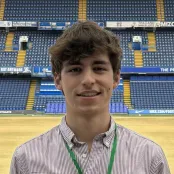 An image of David standing in a large atrium they're using as a night shelter. There a rows of tiered blue seats behind David, who is smiling for the photo.
