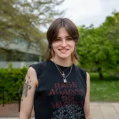 A student stands smiling outside the Physics building dressed casually in a sleeveless band T-shirt.