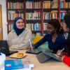 Nora and 2 friends study together in a library. They have their laptops open on the table in front of them.