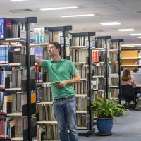 A student selects a book from a shelf while other students work at desks in the background